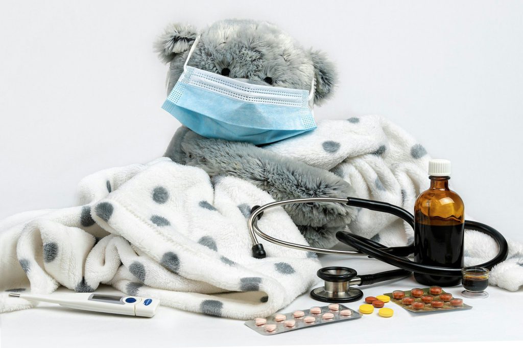 A grey teddy bear is sitting under a polka dot blanket while wearing a blue face mask with a stethoscope and some tablets next to him